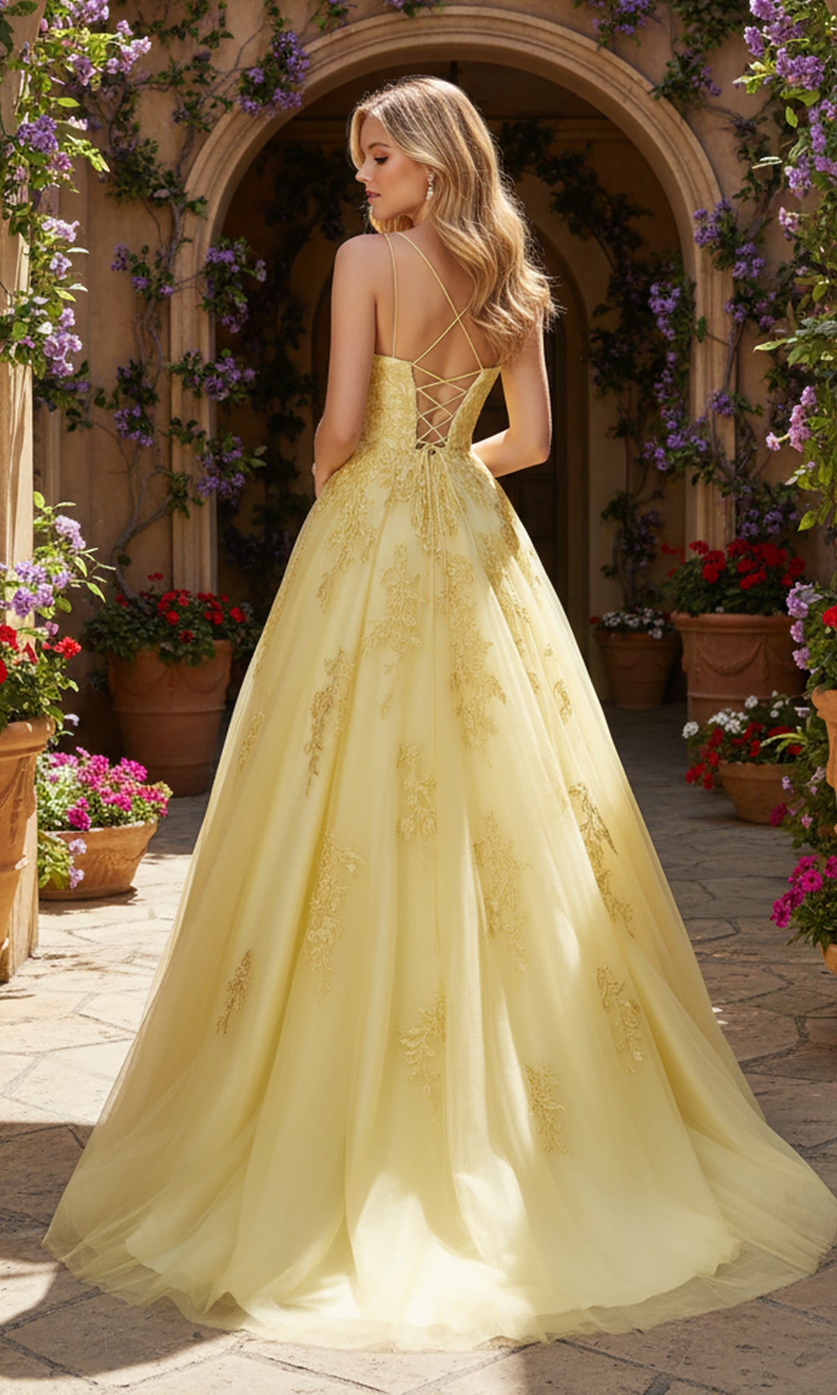 A woman in the PromGirl Exclusive 250228, a yellow floor-length lace prom dress with an adjustable lace-up back, stands outdoors by potted flowers and an arched doorway.